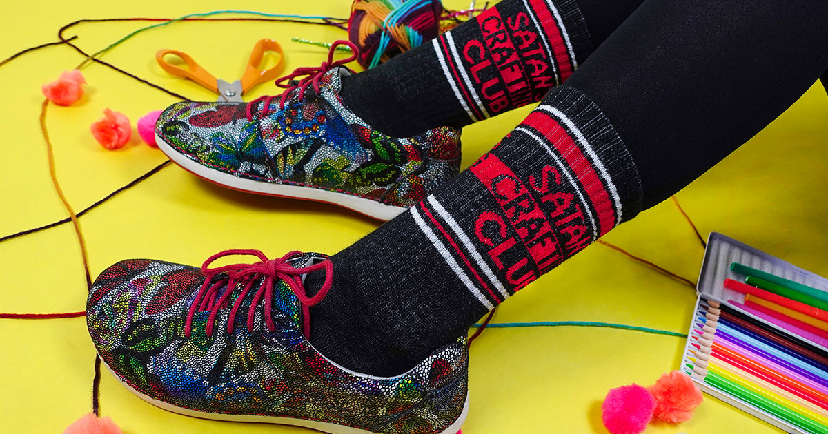 Novelty socks that say "Satan's Crafting Club" on the side are modeled by a woman surrounded by arts and crafts supplies.