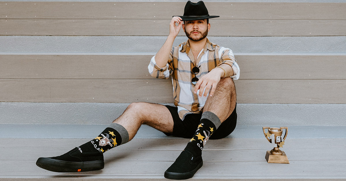 A man wearing GOAT, Greatest of All Time men's socks, sits on a staircase beside a gold trophy,