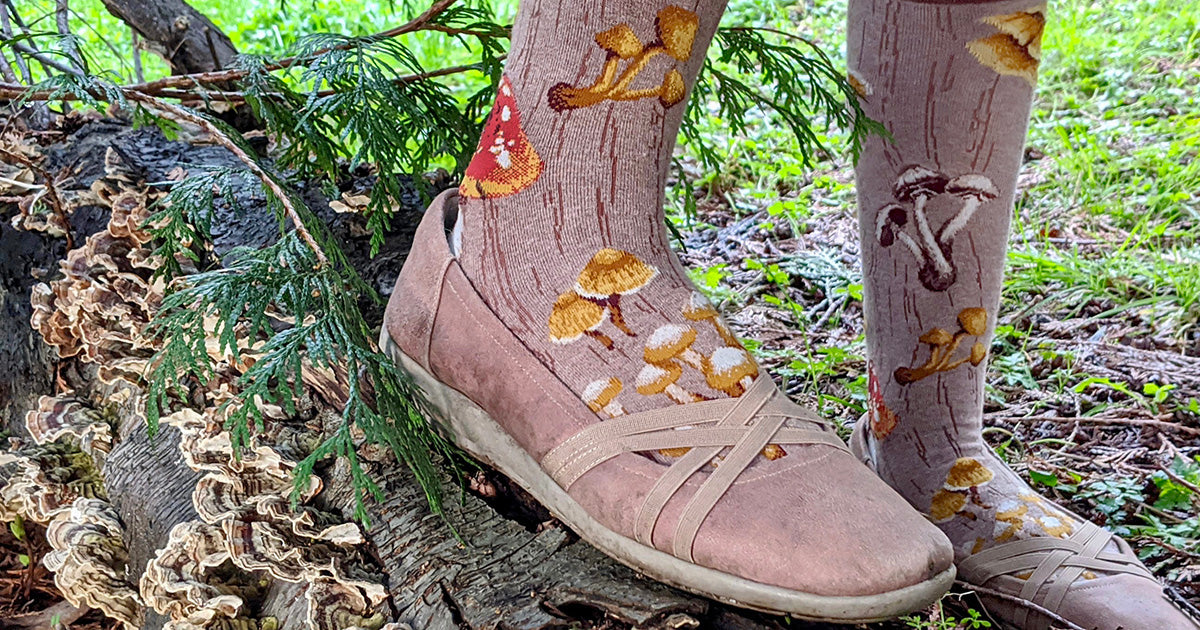 Feet in brown mushroom novelty socks and matching shoes stand on a log covered in small mushrooms