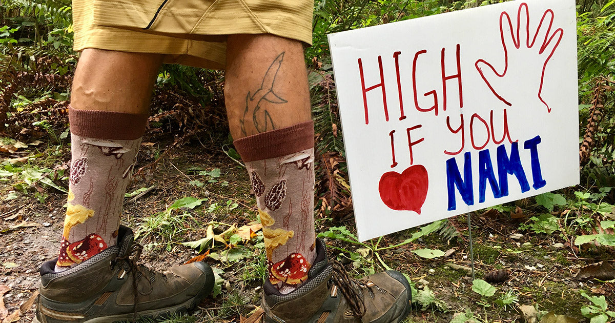 A man wears mushroom socks next to a sign that says "High 5 if You Heart NAMI."