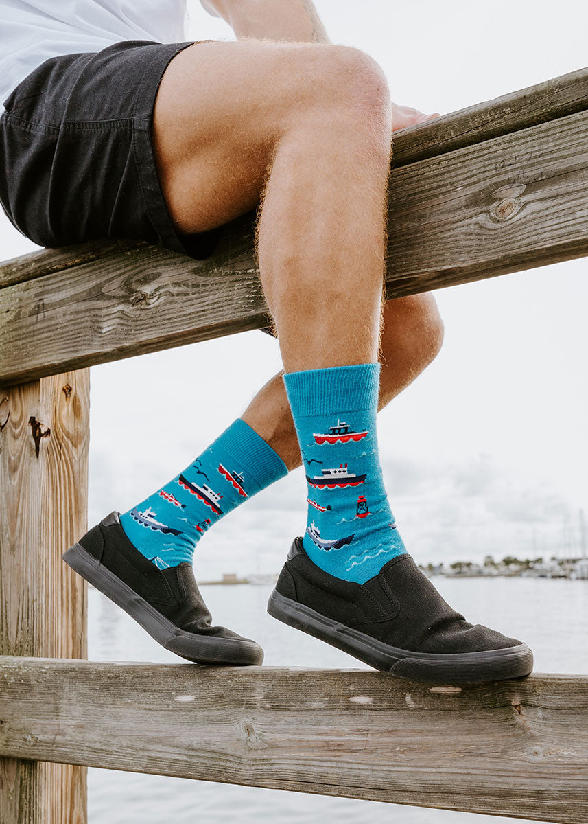 A model wearing boat-themed novelty socks poses sitting on the edge of a pier with the ocean in the background.