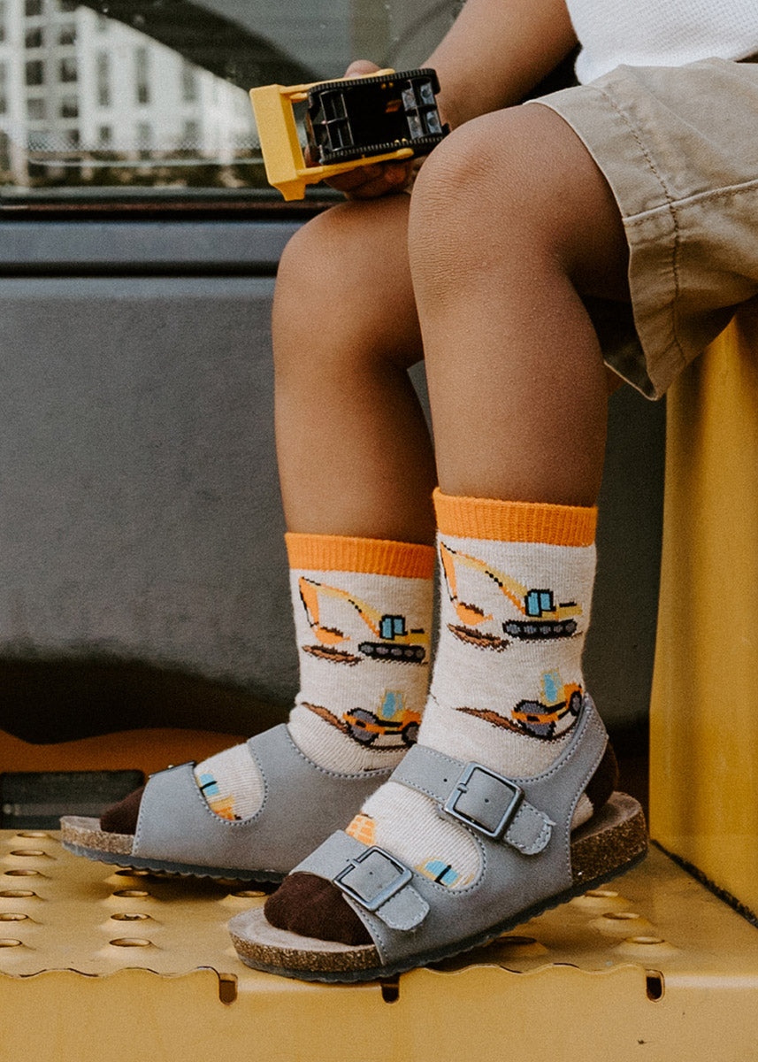 Heavy machinery-themed socks for kids feature dump trucks, steamrollers and backhoes moving piles of dirt on a heather cream background.