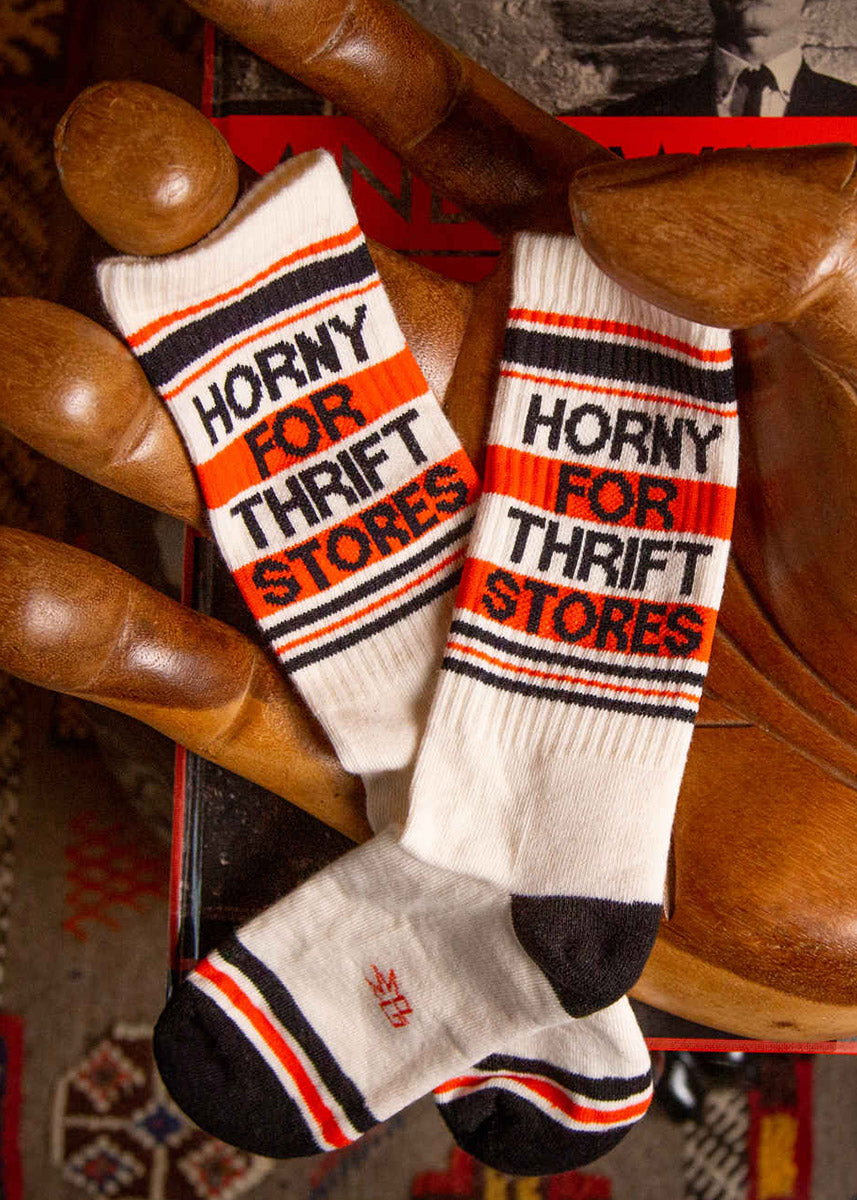  A flat lay of two cream retro gym socks that say “HORNY FOR THRIFT STORES&quot; being held by a large wooden hand.