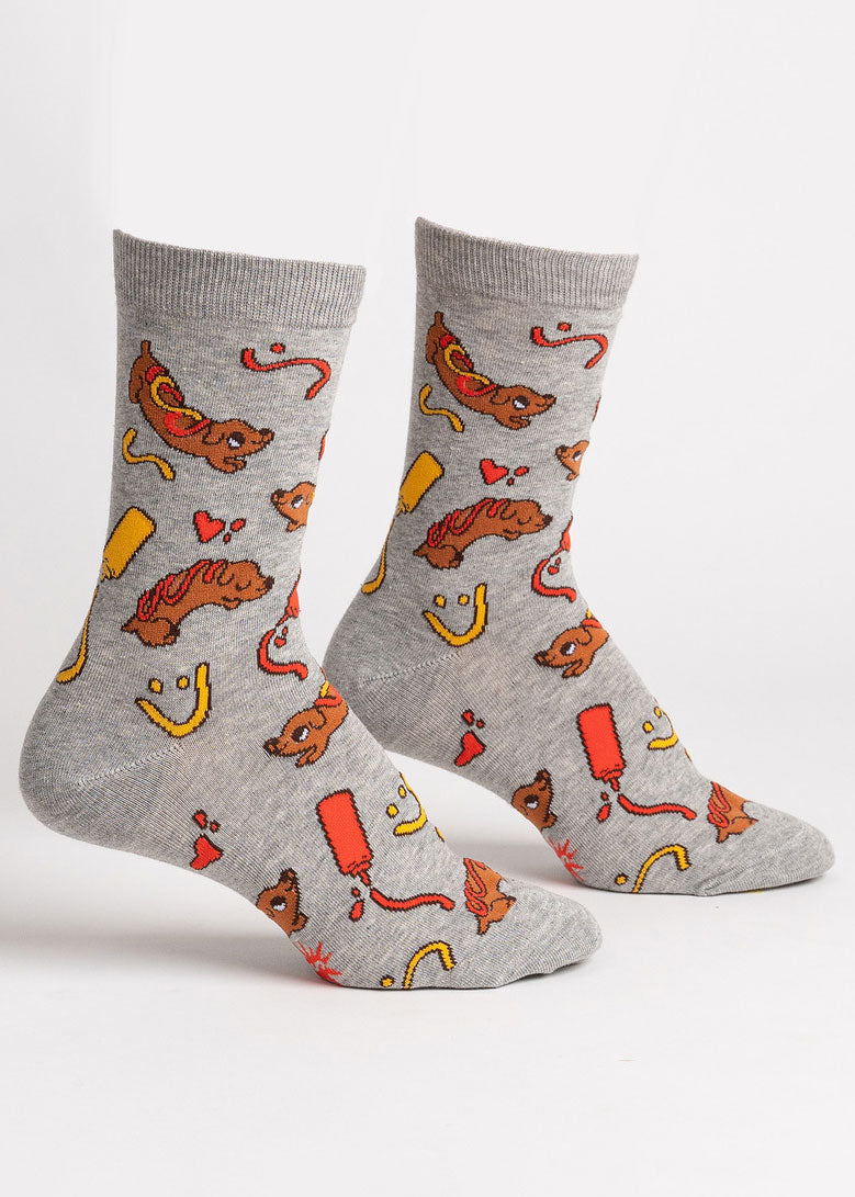 Gray socks for women feature a repeating pattern of brown hot dog-shaped dachshunds, with yellow mustard and red ketchup designs drizzled on their backs and in fun shapes around them, such as hearts and smiling faces. 