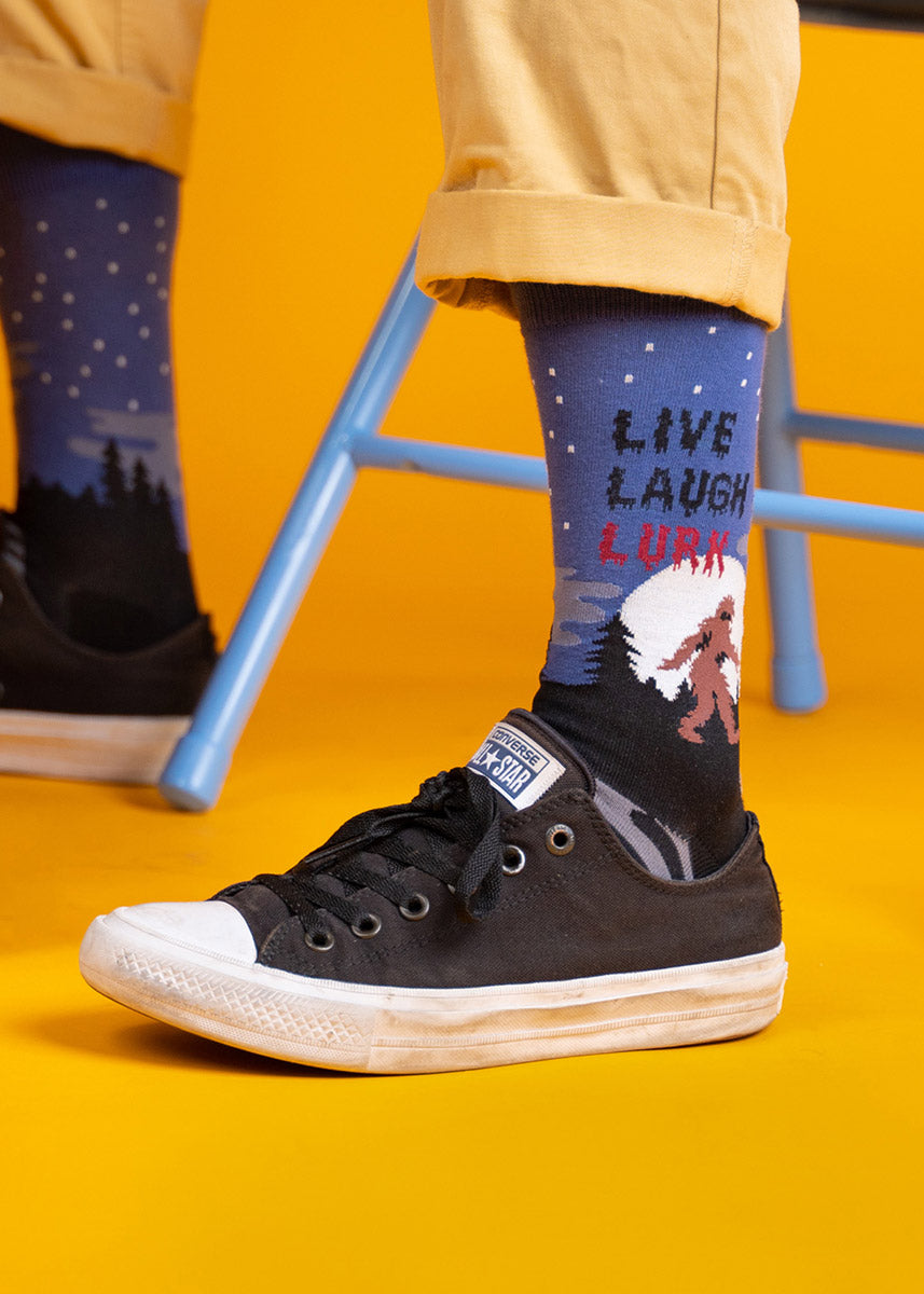 A model wearing bigfoot-themed novelty socks that read &quot;Live Laugh Lurk&quot; poses against an orange background.