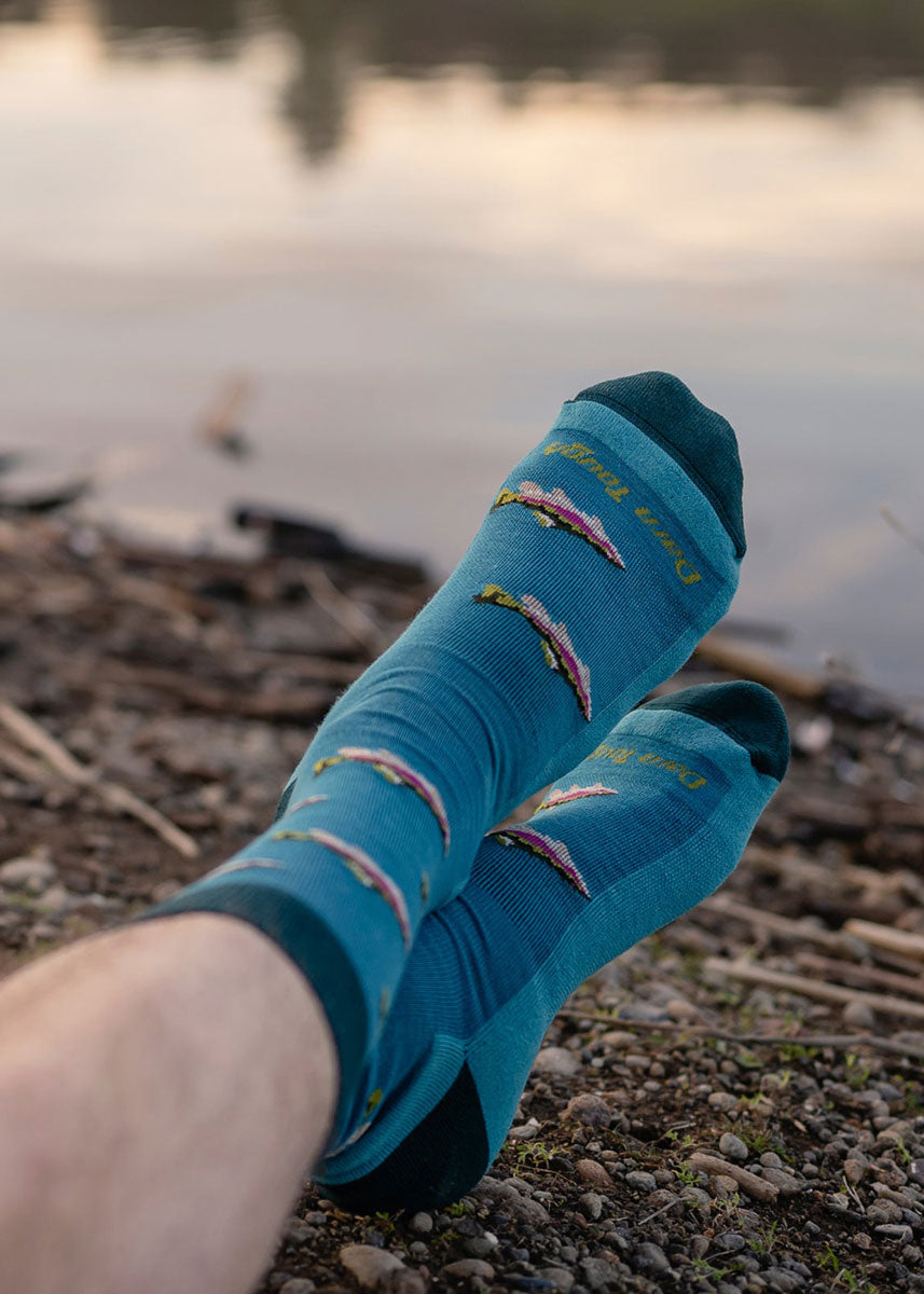 A model poses with their feet crossed by a body of water wearing blue fish-themed wool socks.
