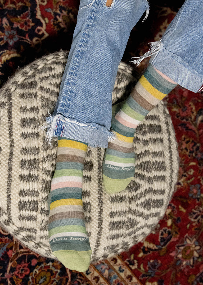 A model wearing green, yellow, white, and pink striped wool socks poses with their feet propped up on an ottoman.