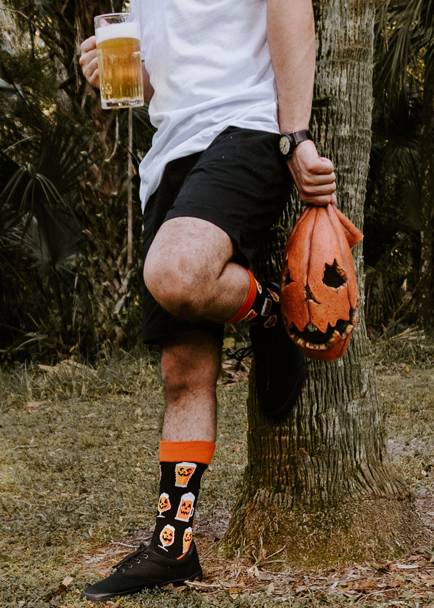 A male model wearing pumpkin beer-themed novelty socks poses against a tree holding a pint of beer and a jack-o-lantern mask.