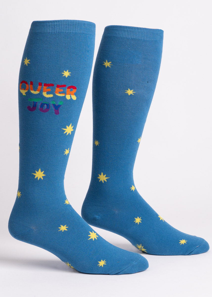 Blue wide-calf knee high socks with an allover pattern of yellow stars and the words "Queer Joy" in rainbow lettering. 