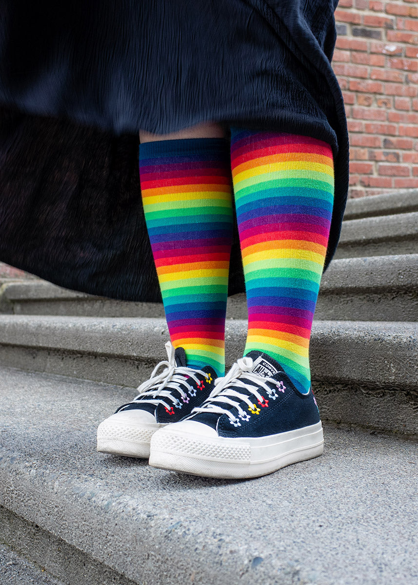 A female model wearing rainbow gradient knee highs and a long black skirt poses outside on stone steps.