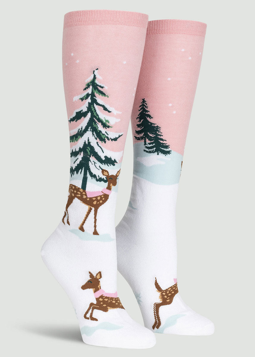 Holiday knee-high socks for women featuring an iridescent white, snowy landscape of pine trees and deer wearing pink scarves against a light pink sky.