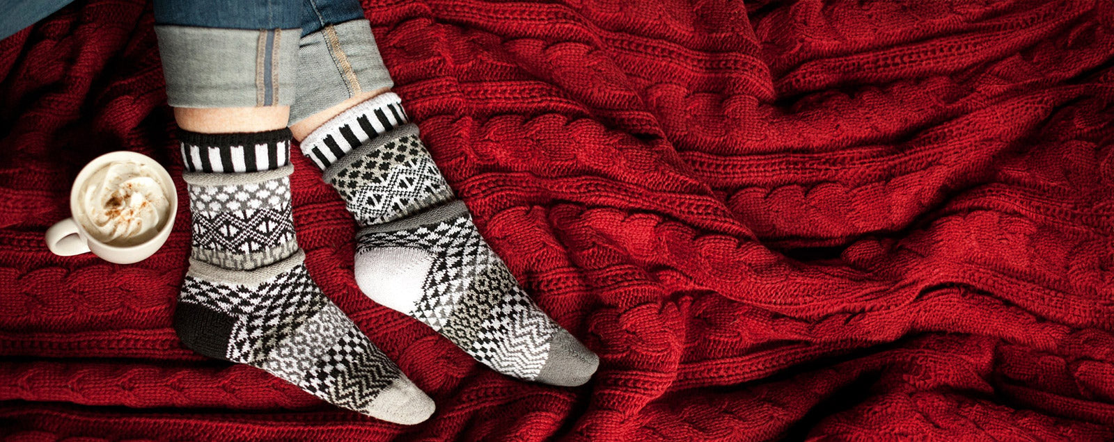 A person wears warm, cozy socks with a black, white and gray pattern while snuggling on a red cable-knit blanket and drinking cocoa.