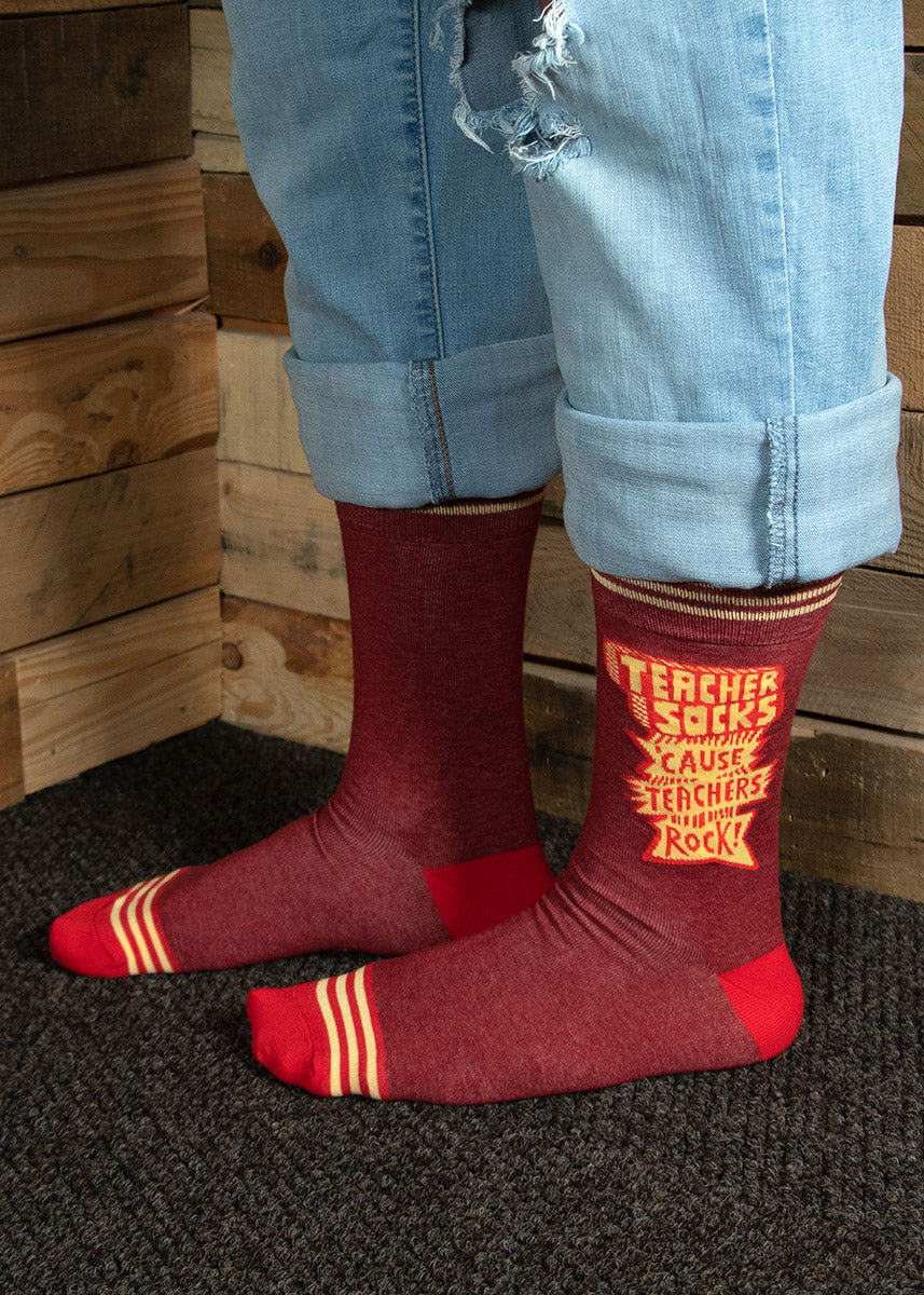 A model wearing novelty socks that say &quot;Teacher Socks &#39;Cause Teachers Rock!&quot; and cuffed jeans poses indoors. 