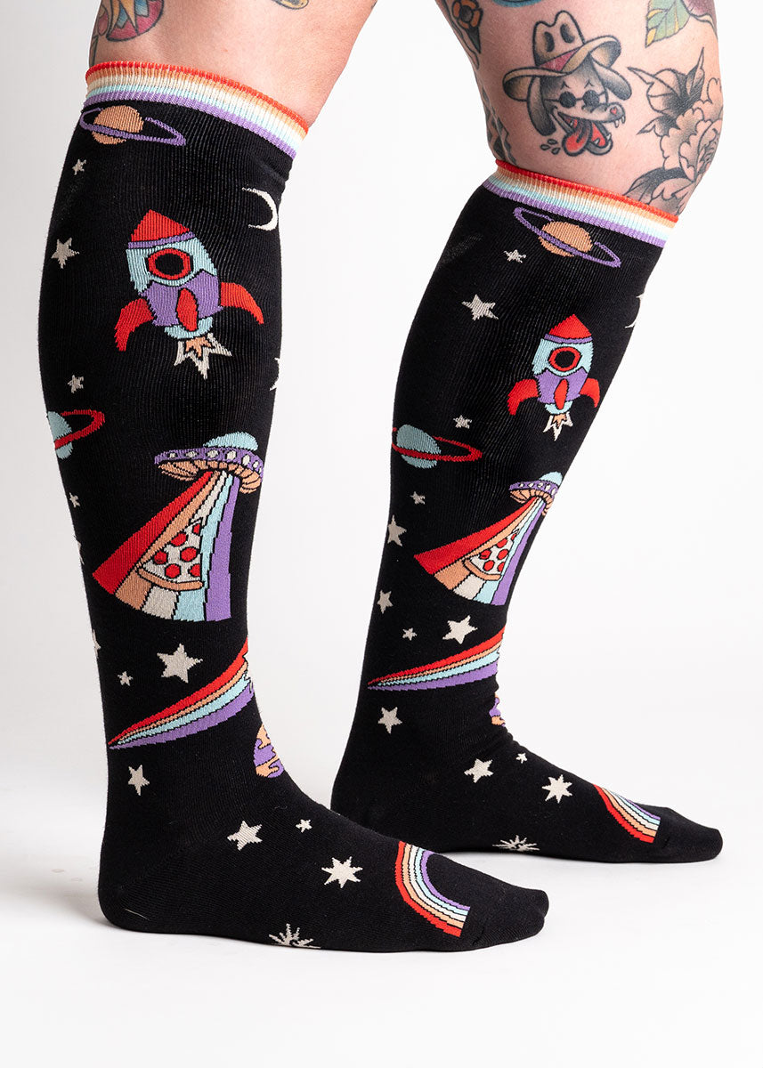 Black space-themed knee-high socks feature stars, planets, rainbows, spaceships and a UFO beaming up a slice of pizza. 