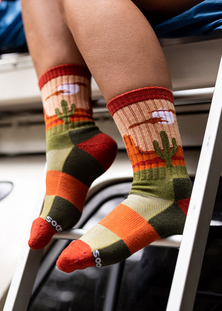 Red, orange, and green wool hiking socks for women feature a desert landscape scene with cacti and sandy dunes.