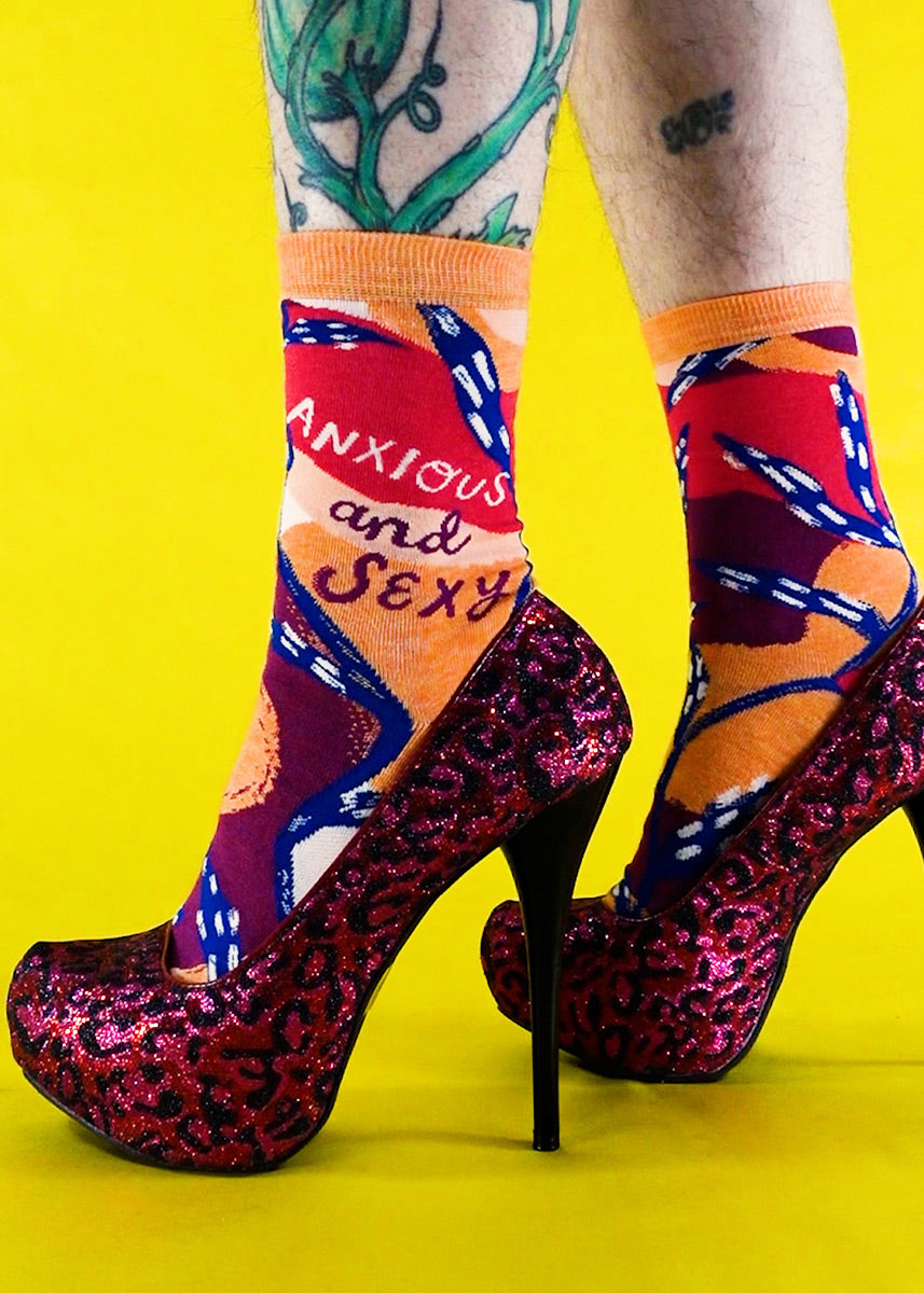 Funny socks for women say "Anxious and Sexy" over a bold abstract background of red, orange, burgundy, and dark blue.