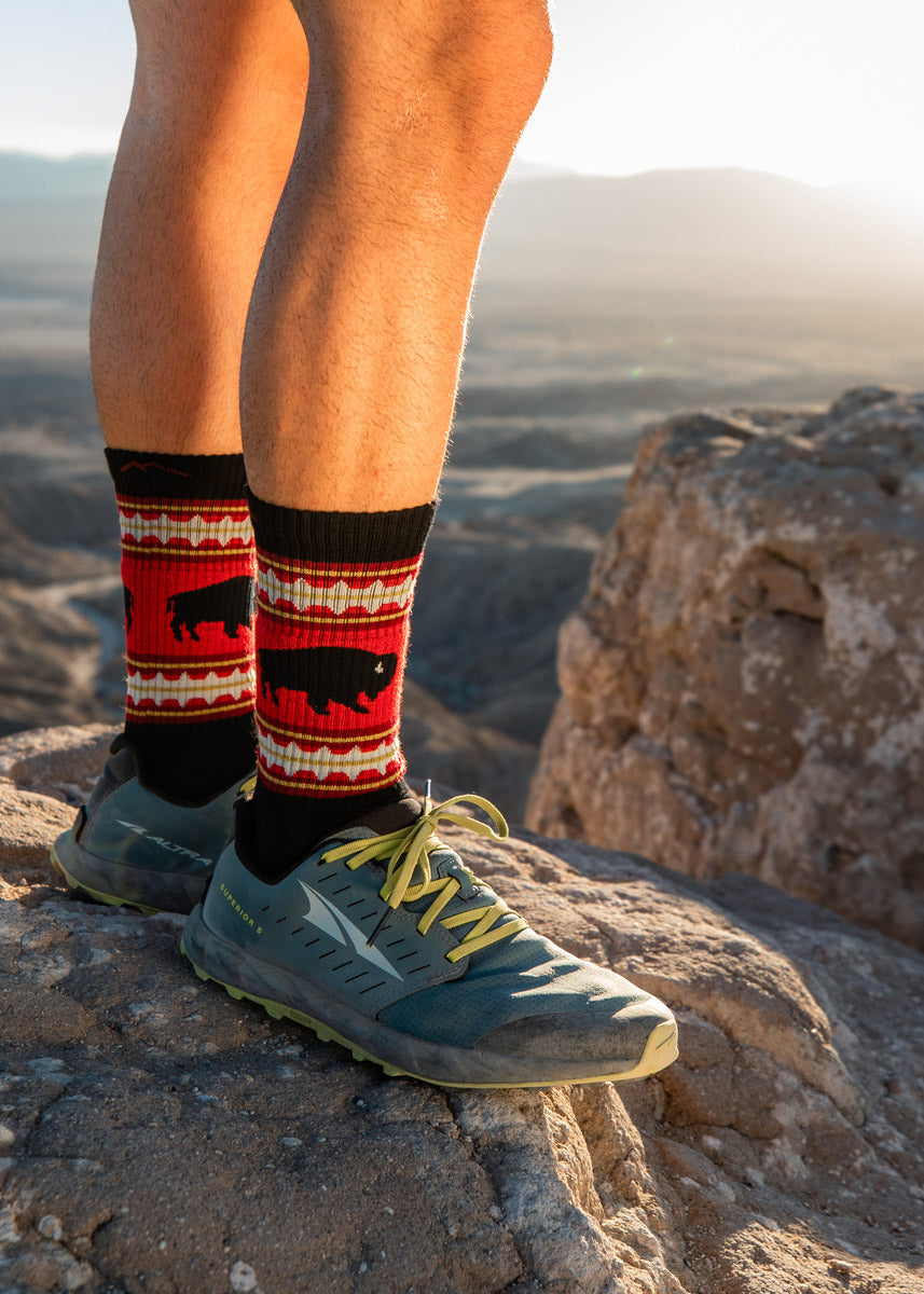 A model wearing buffalo-themed hiking socks poses outside on a cliffside. 
