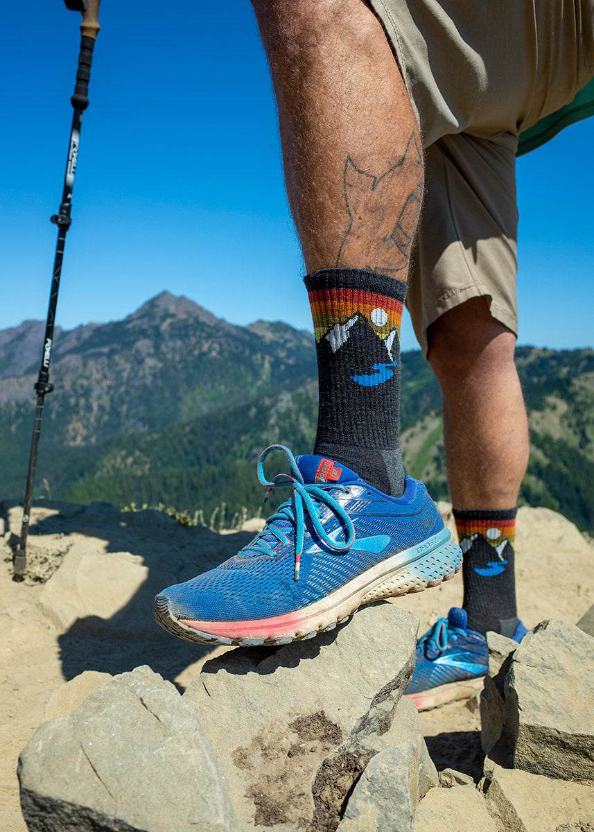 Wool hiking socks for men feature a design of a mountain and a lake under a setting sun and feature footbed cushioning for extra comfort.