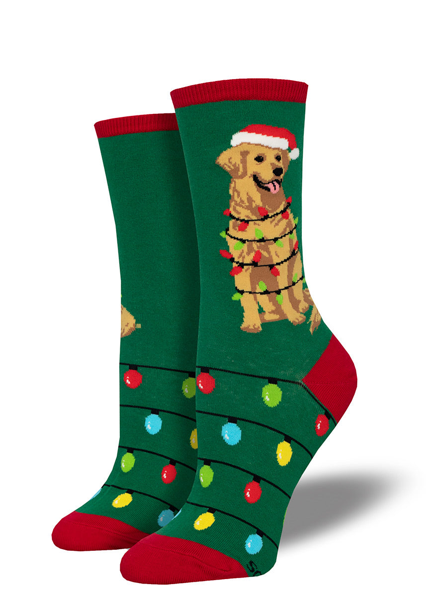 Green women's crew socks with red accents show a golden retriever dog wearing a Santa hat while wrapped in a string of Christmas lights on the leg, with additional multicolored lights adorning the foot.