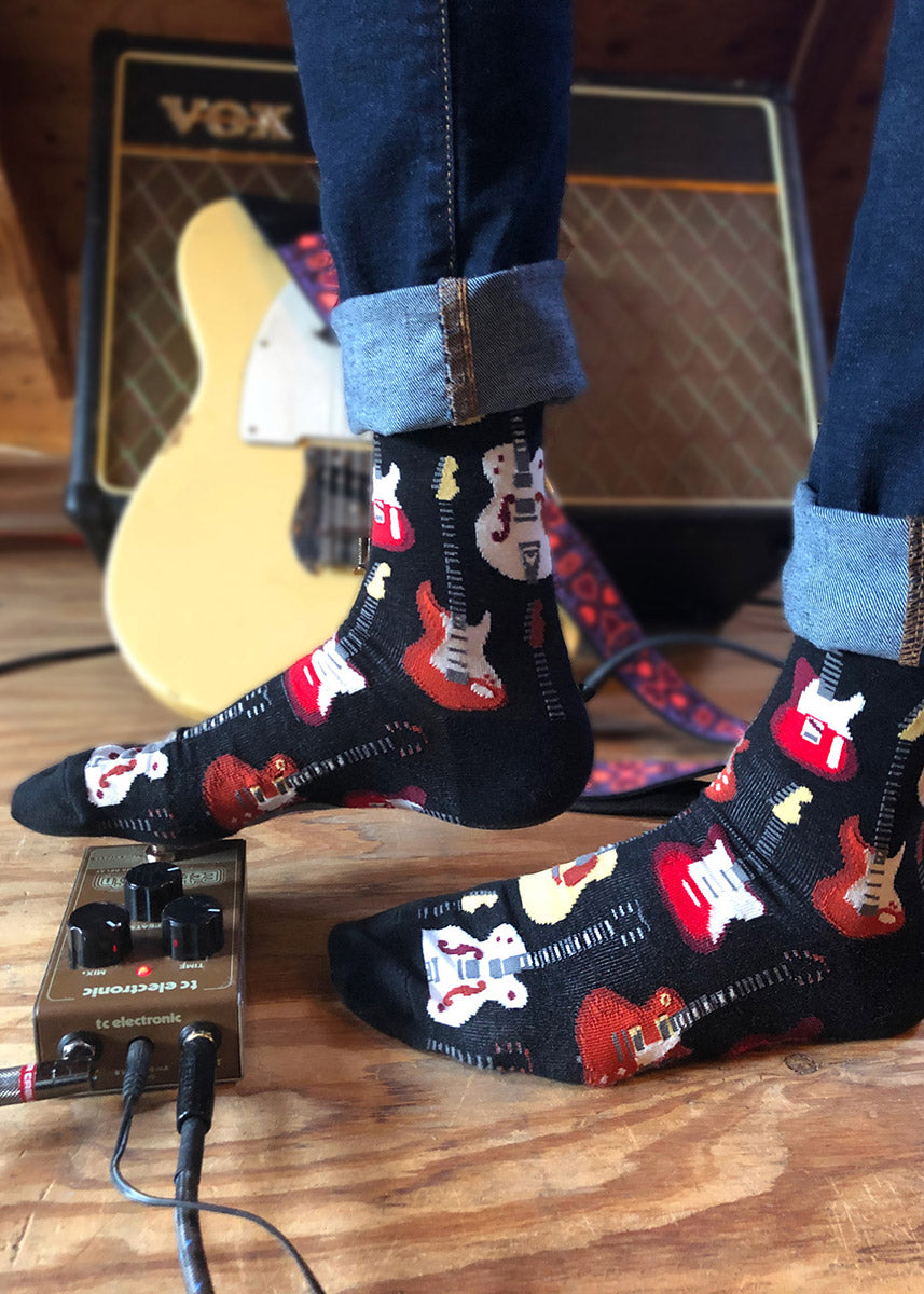 A model wearing guitar-themed novelty socks poses shoeless by a guitar and a guitar amp.
