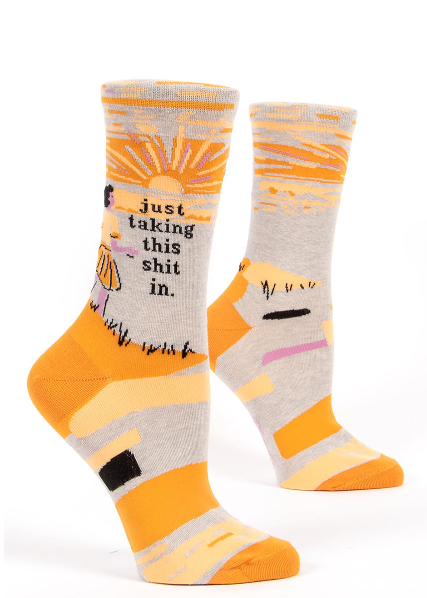 Funny socks for women show a woman enjoying a sunset with the words, "just taking this shit in." 