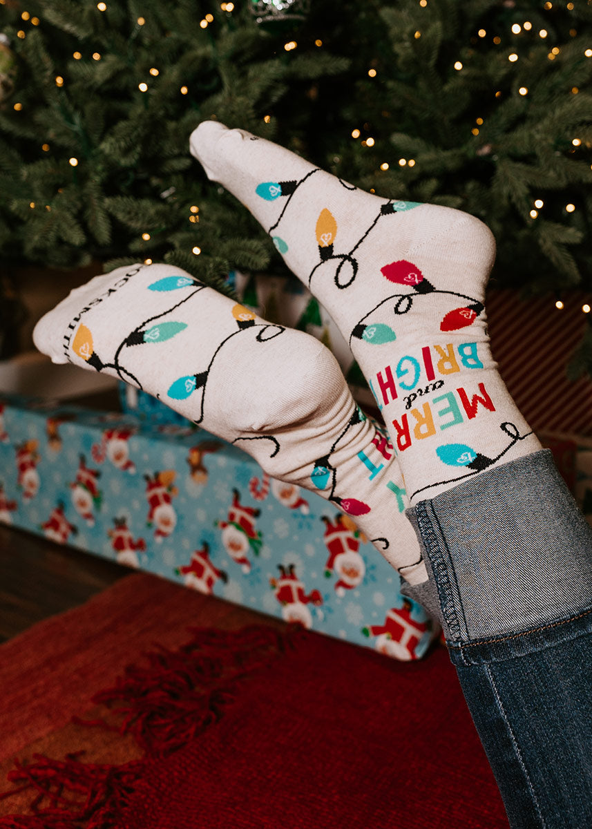 Novelty crew socks for women feature a colorful string of Christmas lights and the words “Merry and Bright” against a heather ivory background.