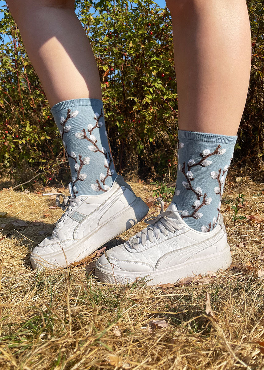 A female model wearing blue pussy willow-themed novelty socks and white sneakers poses outside in a grassy field.