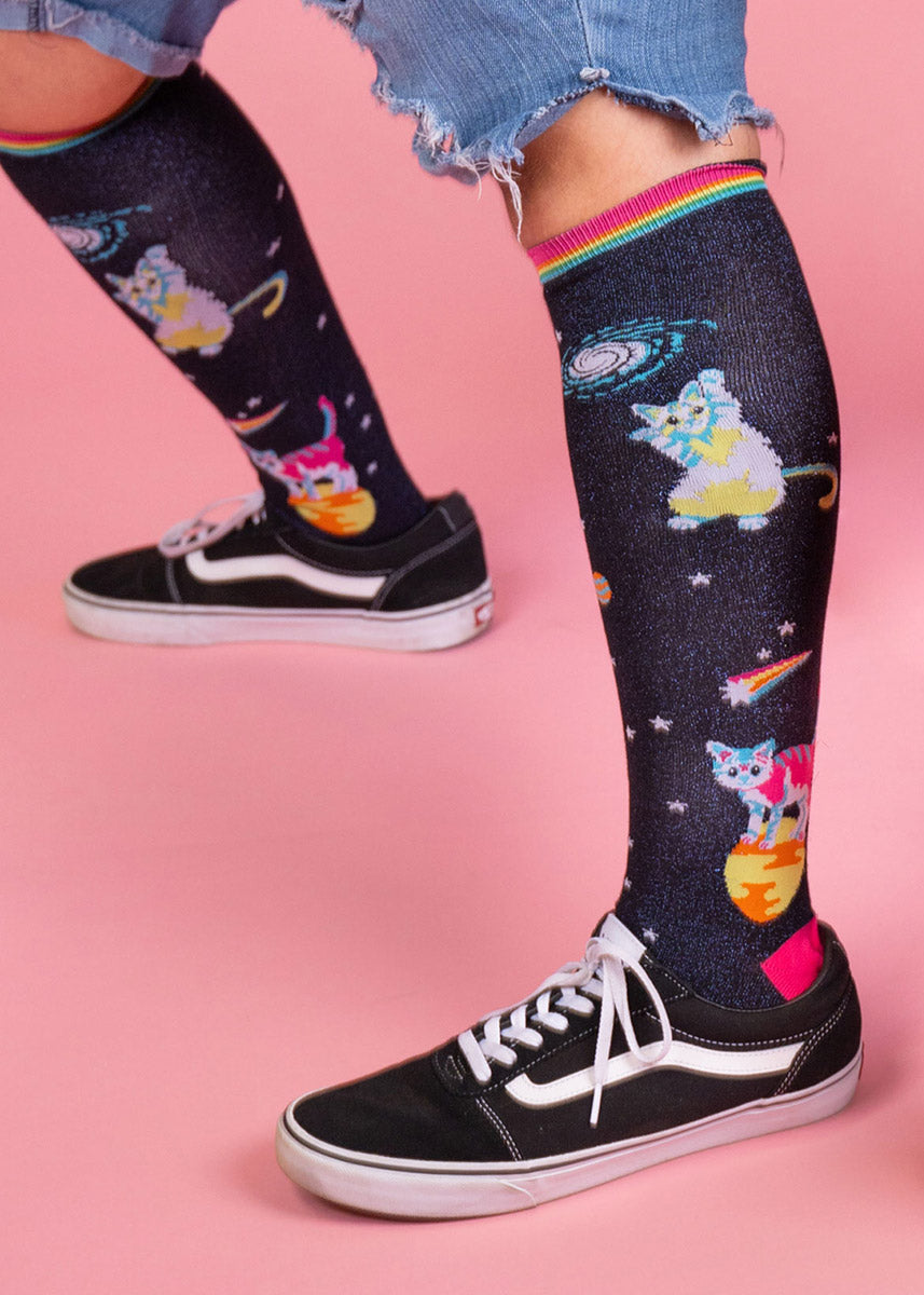 A model wearing space cat-themed novelty knee socks poses wearing black sneakers against a light pink background.