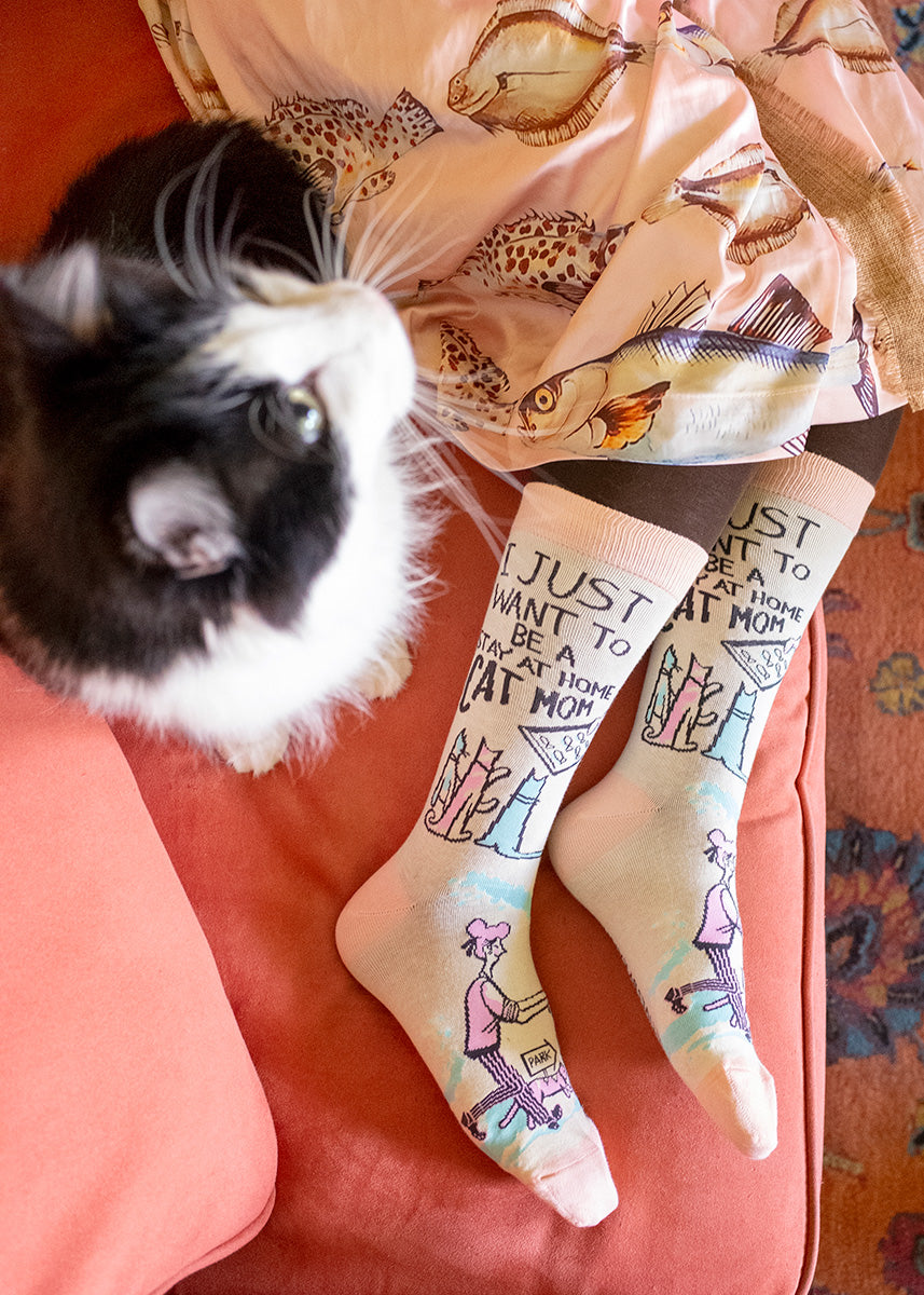A model lays on a coral pink couch next to a black and white fluffy cat, wearing a pair of socks with a beige background, with multicolored cartoon cats on them, with the saying &quot; I Just Want To Be A Cat Mom&quot; on the sides in black.