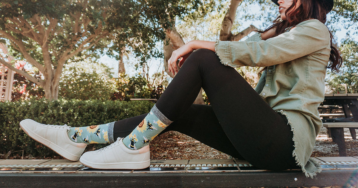 A woman sitting on a bench wears bee and sunflower crew socks with black leggings and white shoes