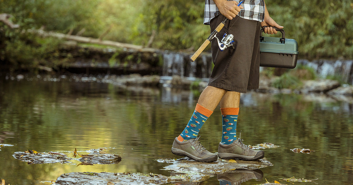 A man in fishing lure socks walks across a river with a fishing pole and tackle box.