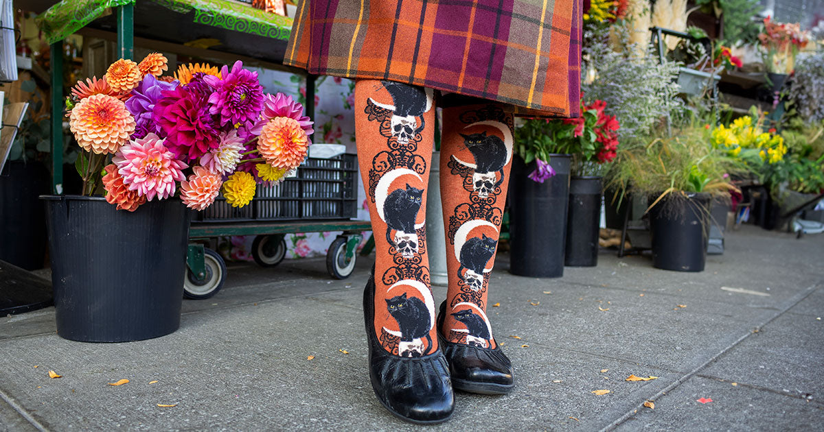 Burnt orange knee socks with a black cat, skull and crescent moon motif worn with a plaid skirt and black shoes beside bouquets of flowers on a sidewalk.