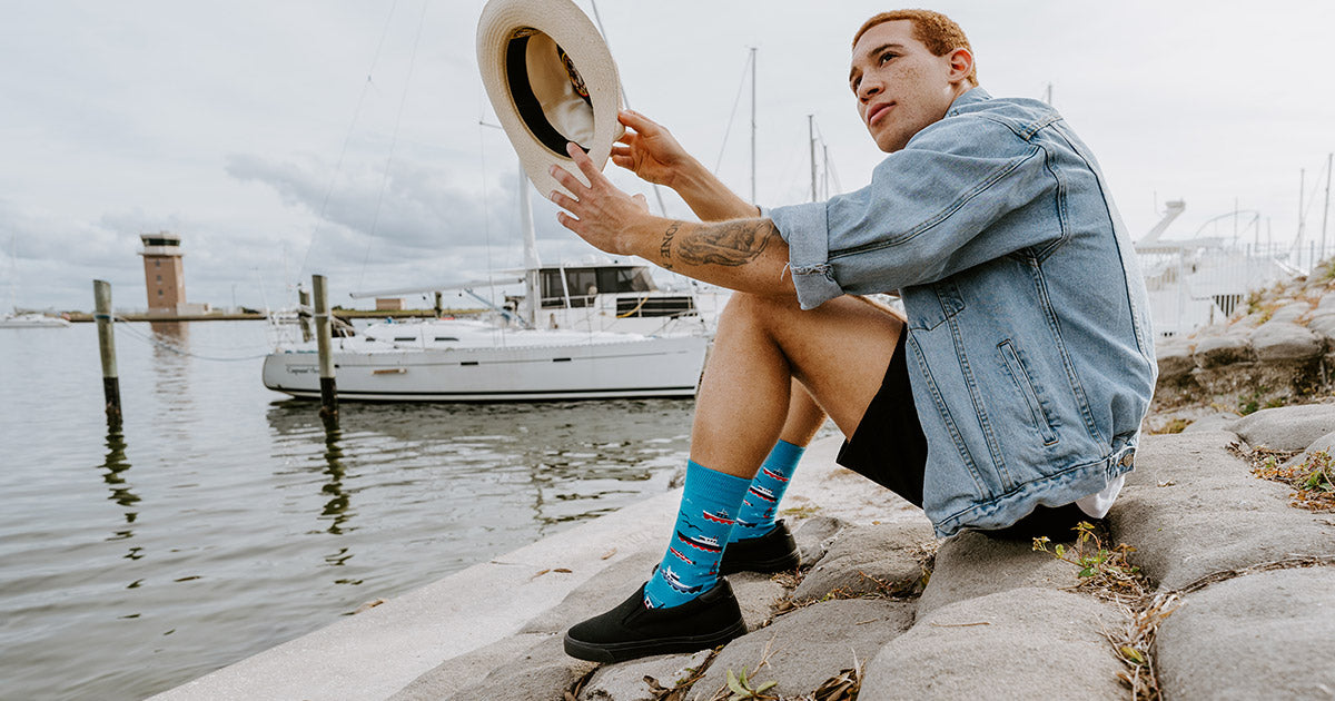 A man wears boat socks with shorts and sits near a pier and a boat on the water