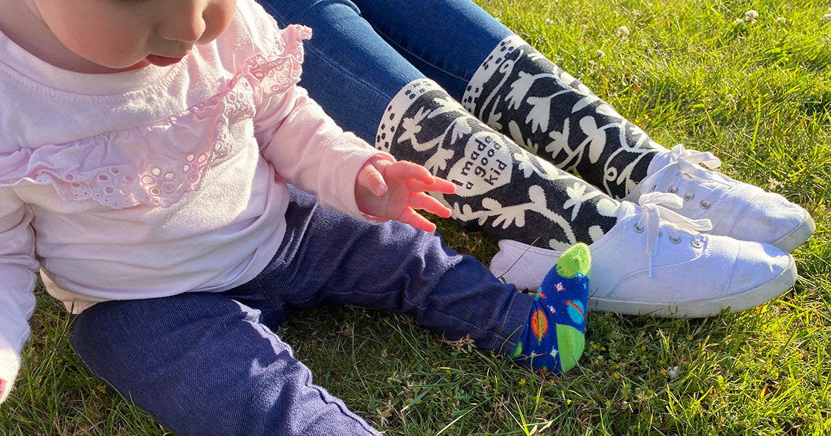 A mom sits beside her young child on the lawn while wearing socks that say "I Made a Good Kid" and white sneakers.
