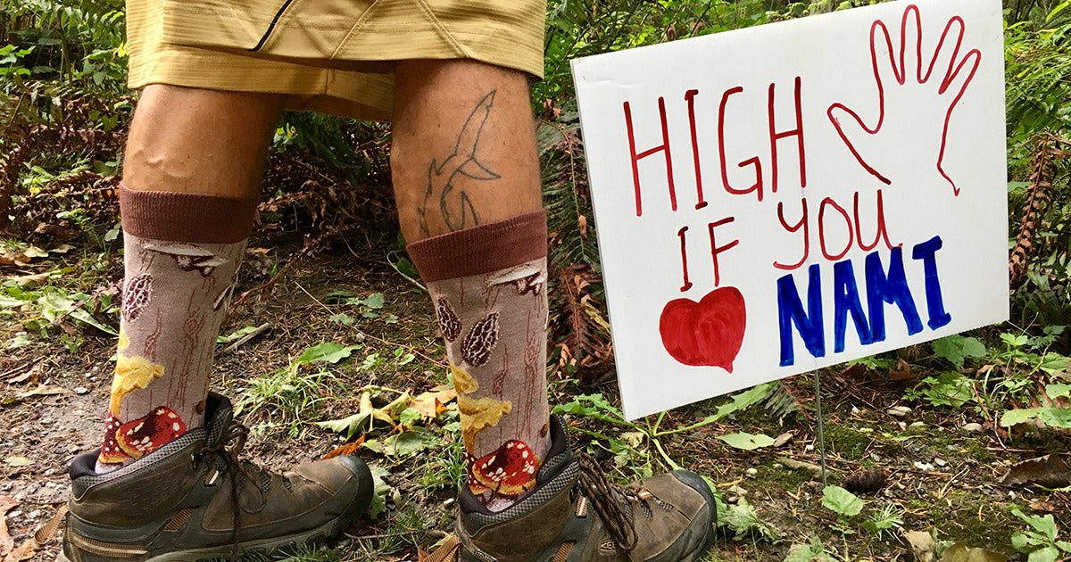 A man wears mushroom socks next to a sign that says "High 5 if You Heart NAMI." 