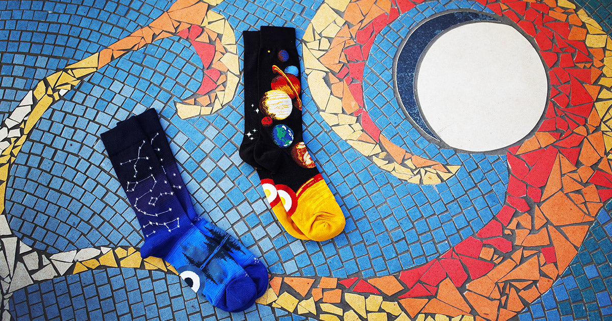 Space socks showing the planets of the solar system and constellations over a striped night sky, with a space-themed mosaic in the background.
