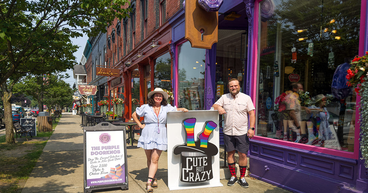 Rachel Northrup McGee and Scotty McGee hold the 'Cute But Crazy' sock store sign while standing in front of The Purple Doorknob sock shop in Ellicottville, New York.