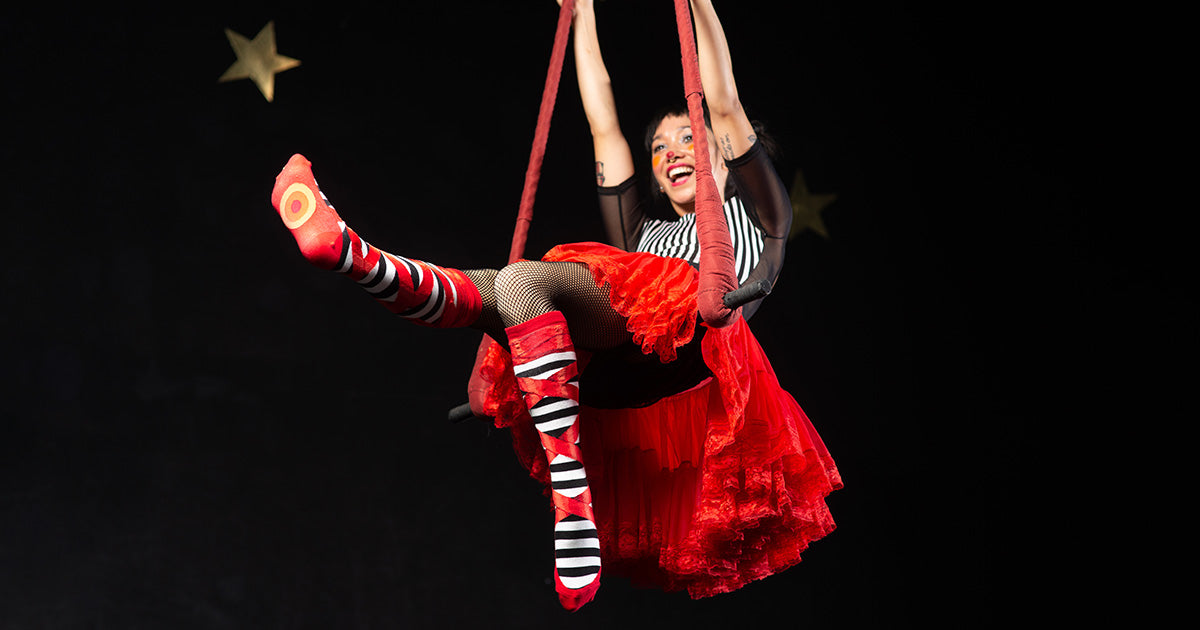 A trapeze performer swings while wearing red faux ballet slipper knee socks with black and white stripes.
