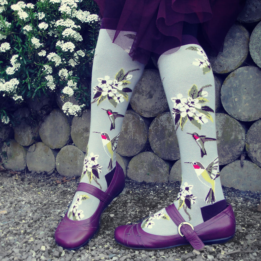 Our Bird Socks collection is the perfect pit stop when shopping for the bird lover in your life. These grey knee highs feature colorful hummingbirds flying to sip nectar from fresh white flowers and have a cute purple heel, toe, and cuff.