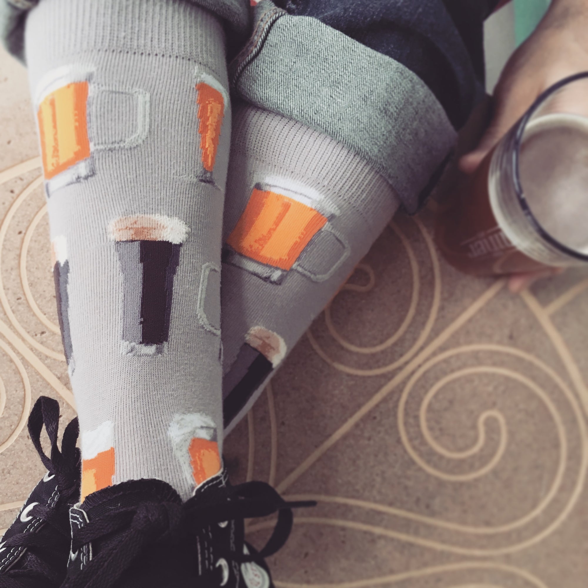 Beer socks featuring all sorts of brew on a gray background are worn by a beer enthusiast as he kicks back and drinks an IPA.