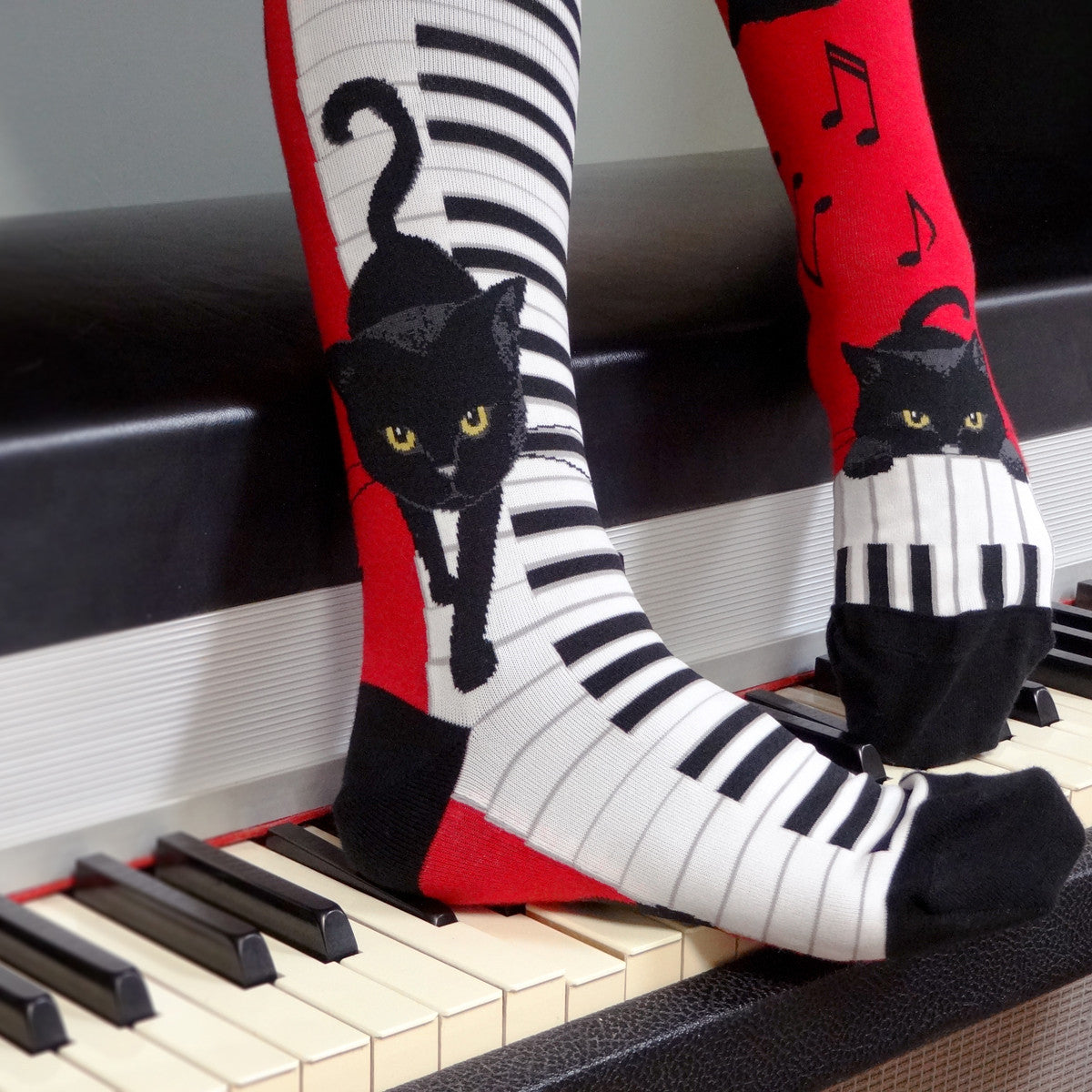 A black cat walks down piano keys while other cats play with music notes on these red knee socks for the music lover designed by sock brand ModSocks.