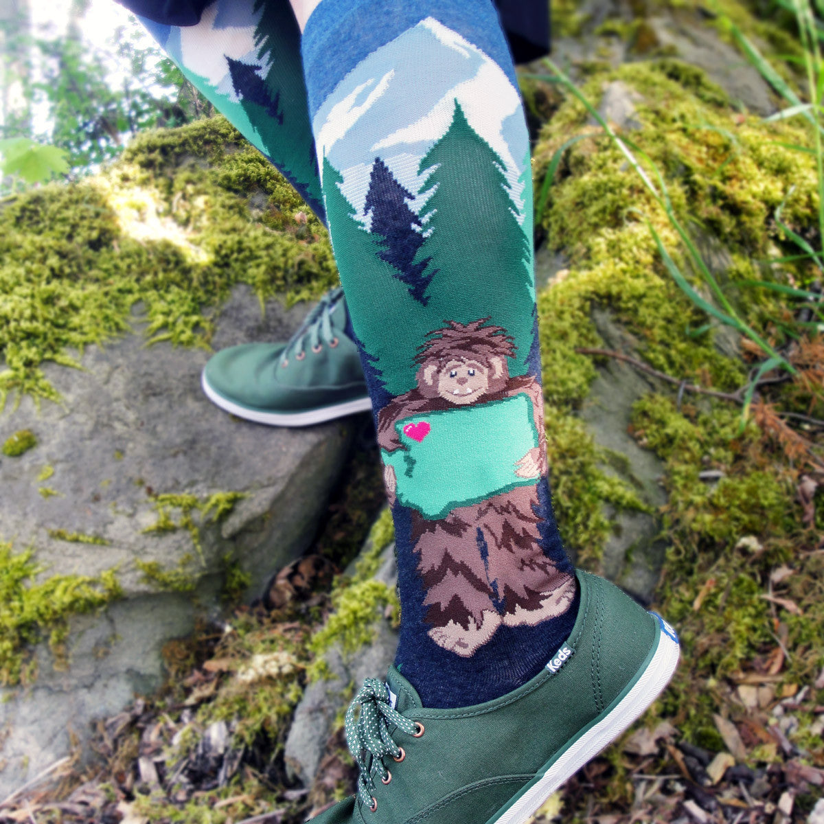 Sasquatch gives the state of Washington a bigfoot-sized hug while standing in front of a mountains-and-evergreen scene on these green and blue knee socks by ModSocks.