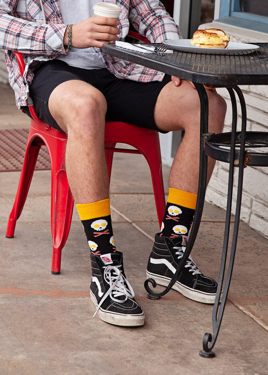 A male model wearing breakfast-themed novelty socks and black Vans poses with a breakfast sandwich at an outdoor cafe.