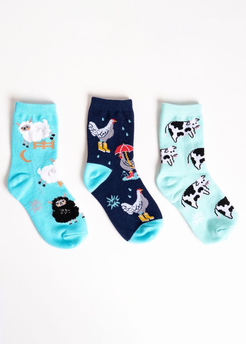 Three pairs of coordinating socks for kids: one pair features fuzzy white and black sheep, another has chickens wearing rainboots and holding umbrellas out in the rain, and the third has black and white spotted cows. 