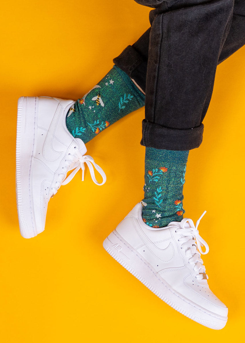 A model wearing bee-themed sparkly novelty socks and white sneakers poses against a yellow background.