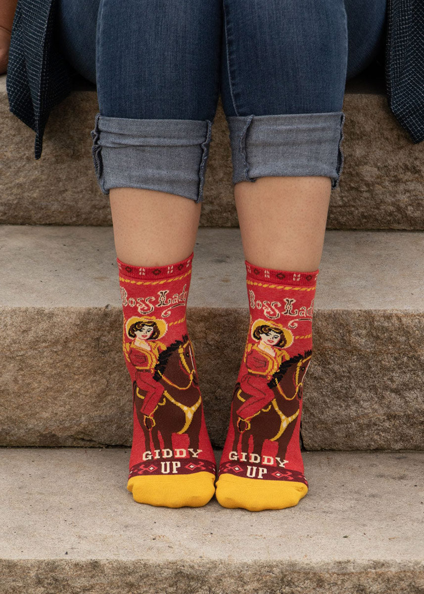 A model wearing cowgirl-themed red novelty ankle socks that read &quot;Boss Lady&quot; poses sitting on stone steps.