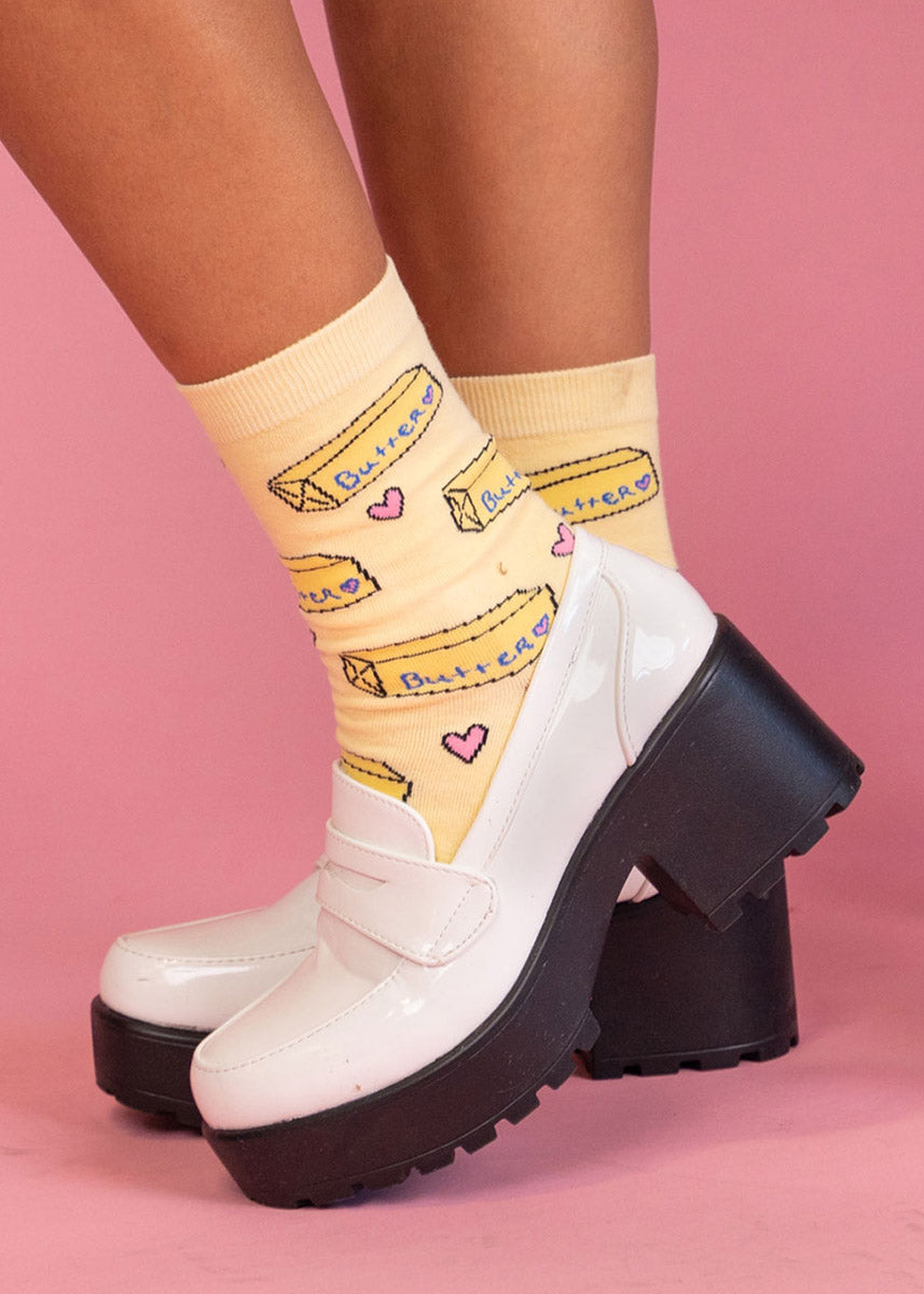 A model wearing butter-themed novelty socks poses against a pink background in white platform shoes.