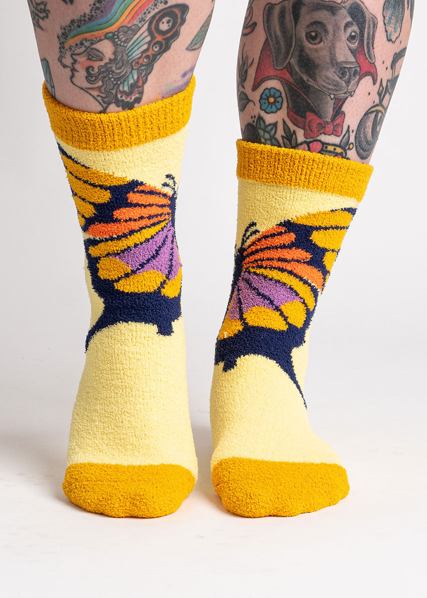 A model with leg tattoos poses against a white background wearing fuzzy yellow slipper socks with a yellow, orange, and purple butterfly on them.