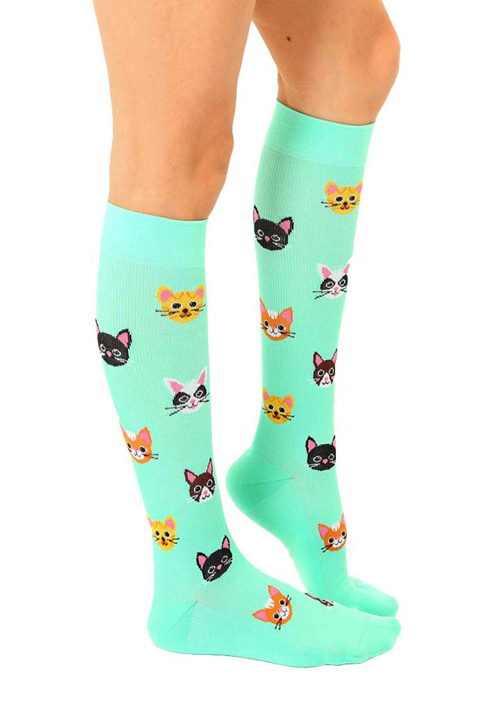 Side view of knee-high compression socks with an allover pattern of various cat faces, including black, orange tabby, tuxedo cats and more.