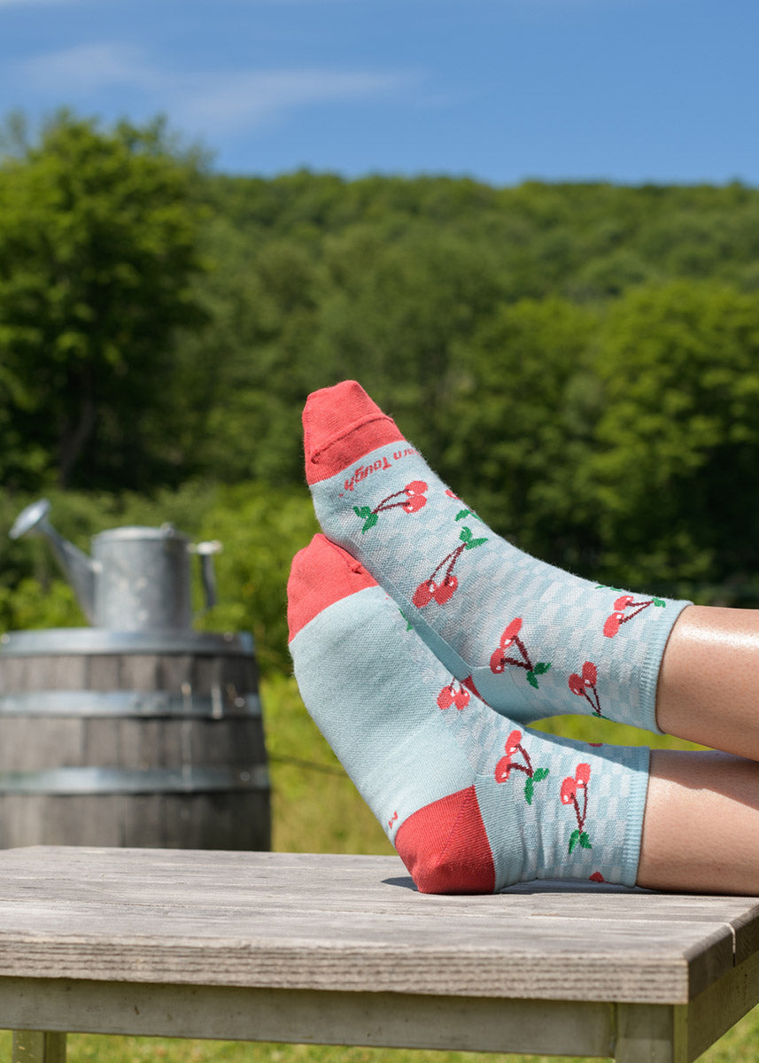 A model wearing cherry-themed wool socks rests their feet on a wooden table against a backdrop of grassy hills and trees.